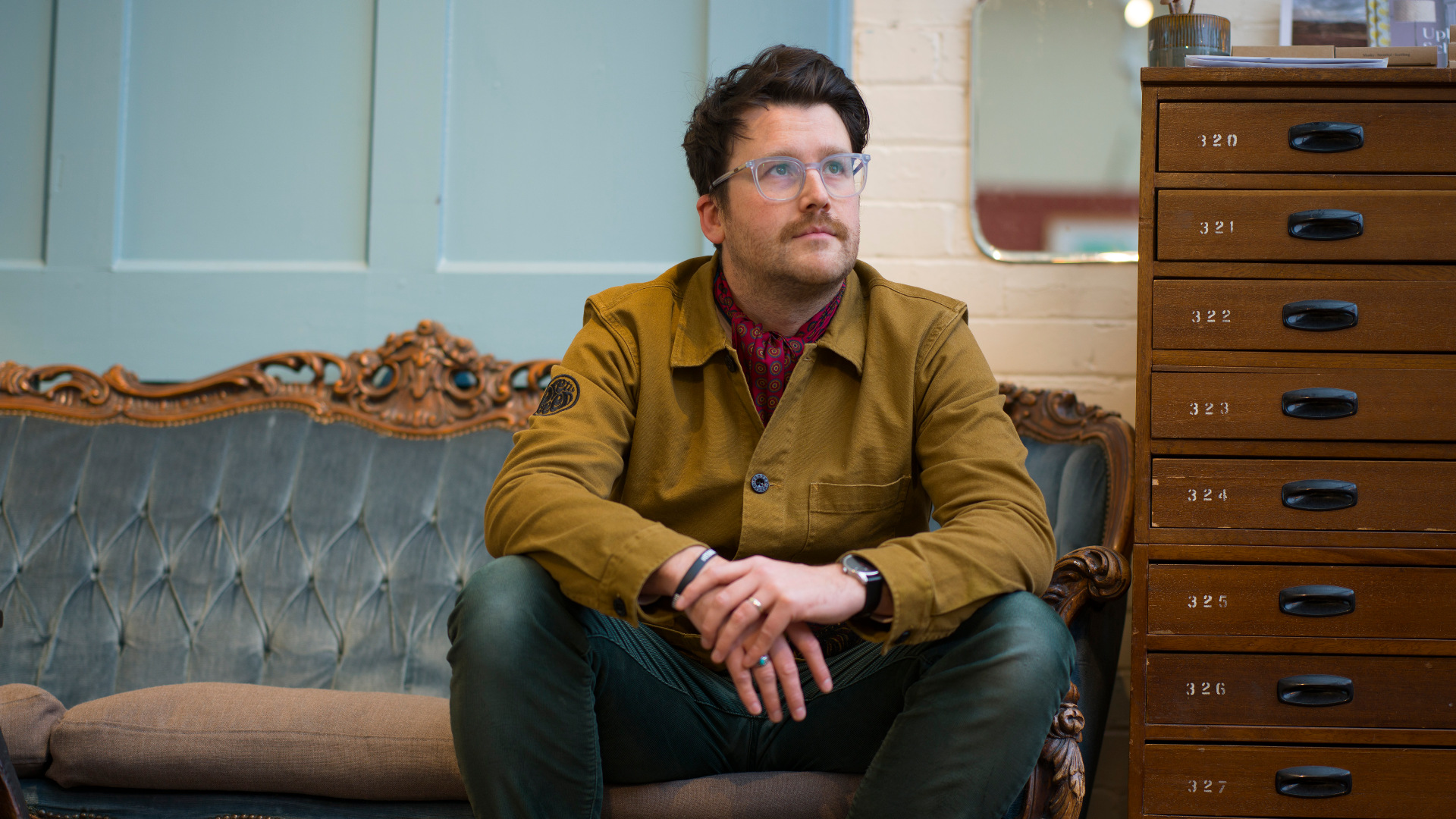 The poet Matt Abbott, sitting on a sofa, looking above and to his left. He wears a brown jacket and clear glasses. Portrait by Stephen Cole.