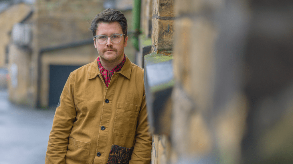 The poet Matt Abbott, standing beside a sandstone mill, looking at the camera. He wears a brown jacket and clear glasses. Portrait by Stephen Cole.